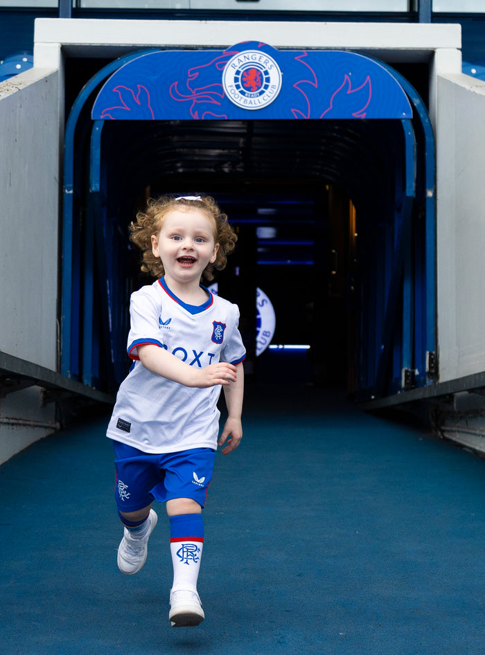 Smiling toddler in Rangers Infant Home Kit runs through a tunnel in a white jersey with crest and blue shorts