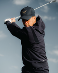 Golfer in midnight navy golf performance hoodie and cap mid swing against a blue sky