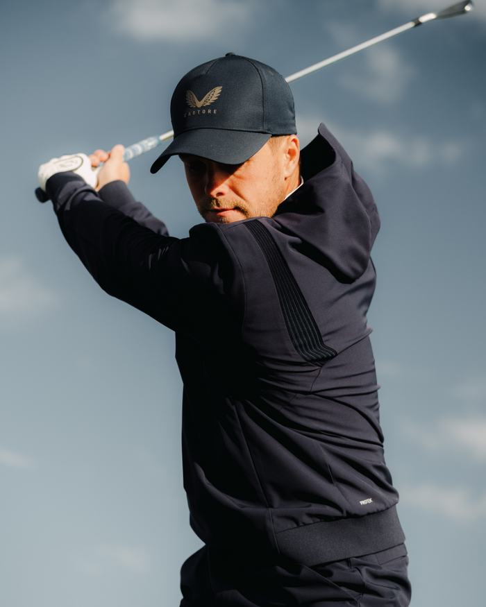 Golfer in midnight navy golf performance hoodie and cap mid swing against a blue sky