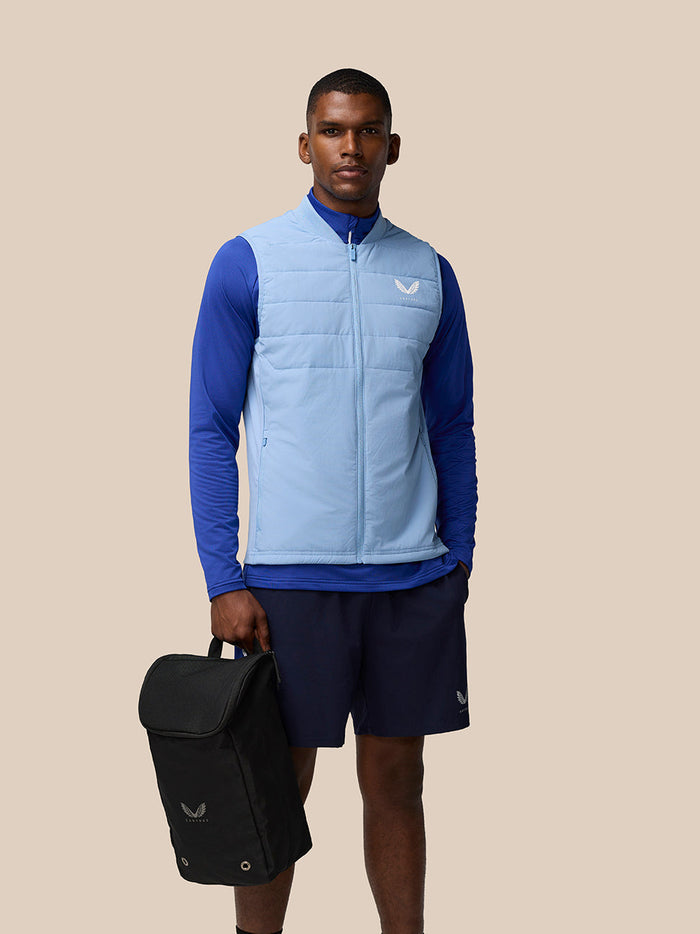 Model wearing a light blue padded vest holds a black boot bag.