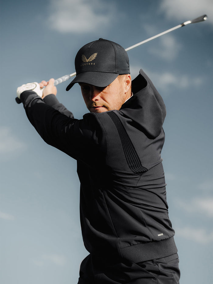 Athlete in black Golf Pinnacle Performance Hoodie with hood up mid swing holding a golf club outdoors