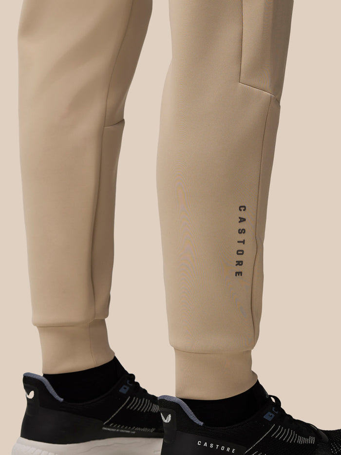 Lower leg view of beige Castore joggers showing the vertical Castore wordmark along the calf and cuffed hem near sneakers