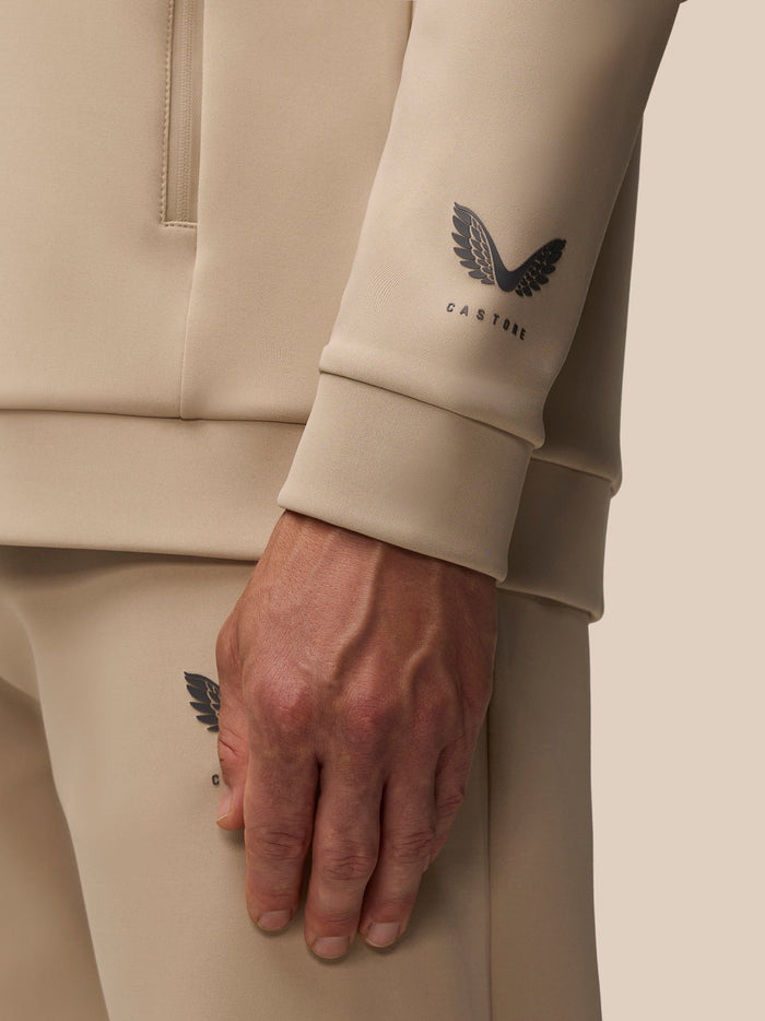 Close-up of the Castore logo on the beige hoodie sleeve with a resting hand.