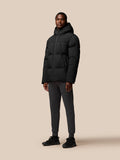 Full-body view of a man in a black puffer jacket with hood down, wearing dark joggers and sneakers