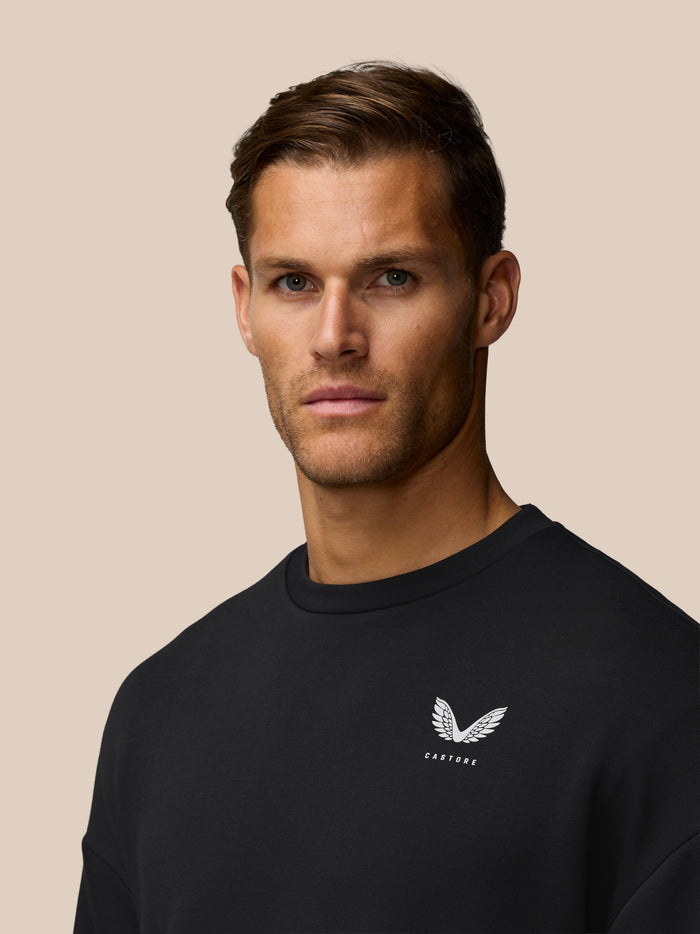 Close up of the black Travel Lifestyle Sweatshirt chest showing a white wing logo and Castore branding.