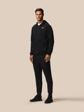 Full body shot of a man in black travel zip hoodie with hood down and left chest logo, paired with matching black joggers.