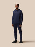 Full-body shot of a man in navy Castore tracksuit with hoodie, standing against a beige background.