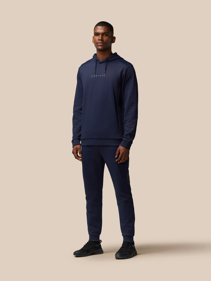 Full-body shot of a man in navy Castore tracksuit with hoodie, standing against a beige background.