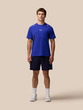 Male model wearing blue Castore T shirt and navy Apex 6 inch shorts with logo on left leg against a beige background