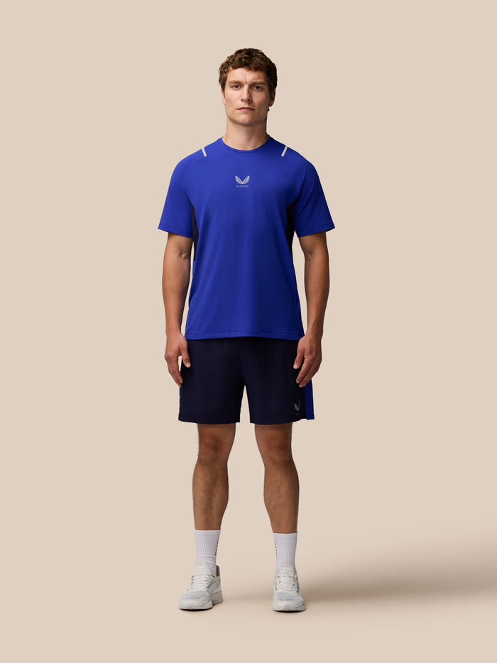 Male model wearing blue Castore T shirt and navy Apex 6 inch shorts with logo on left leg against a beige background