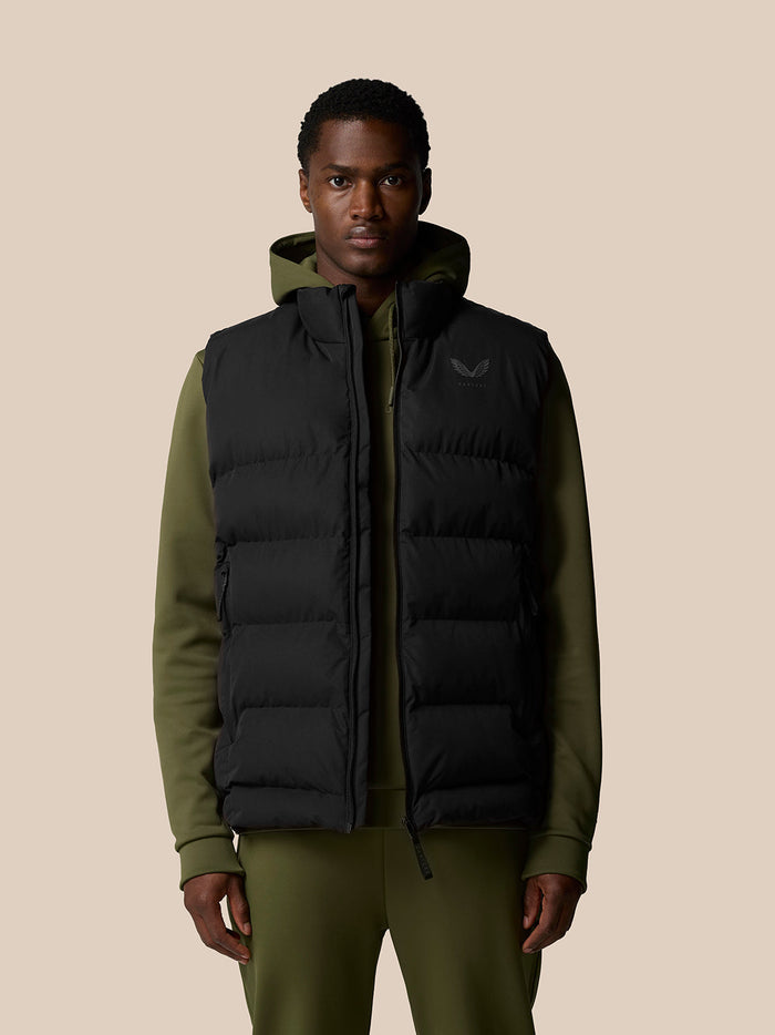 Man wearing black sleeveless puffer gilet over olive green hoodie with front zip.