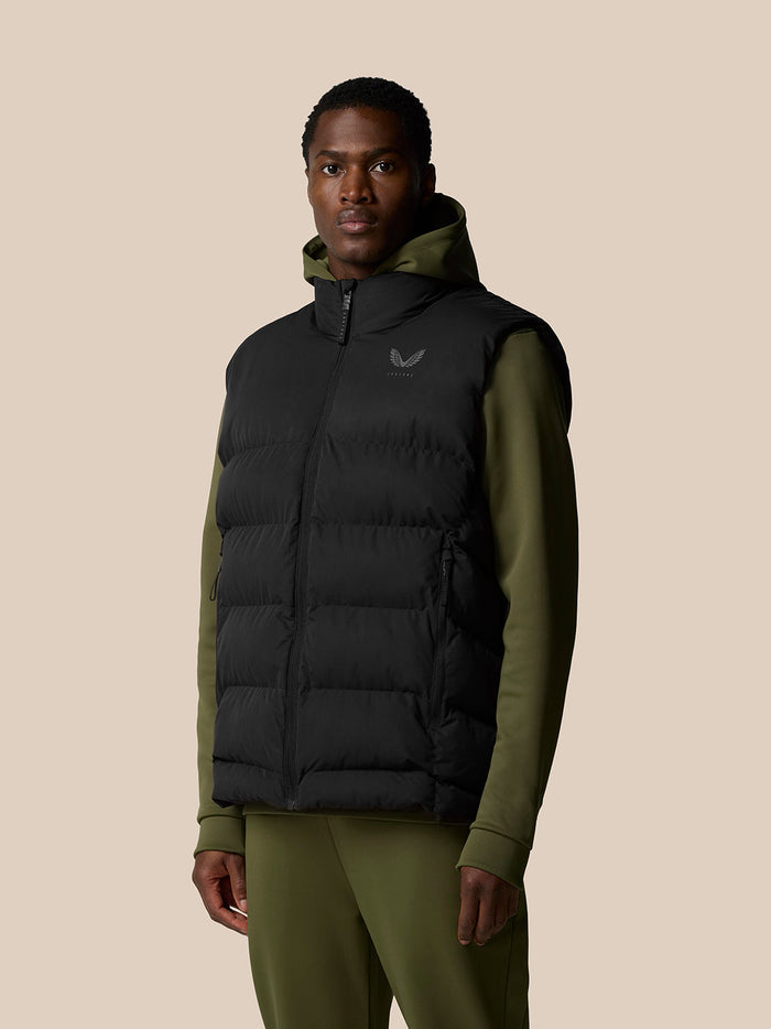 Waist up portrait of a man in a black Puffer Gilet with a hood and olive hood lining, chest logo visible.