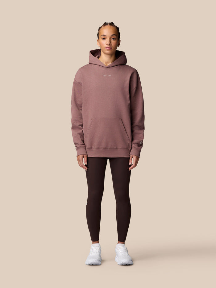 Female model in mauve ADAPT LIFESTYLE HOODIE with hood up and front pocket, full body shot against beige backdrop.