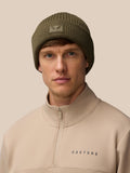 Ivy green ribbed knit beanie with a Castore logo patch on the fold worn by a model against a beige background