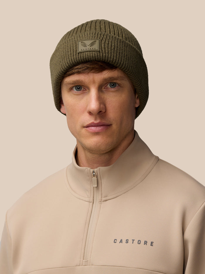 Ivy green ribbed knit beanie with a Castore logo patch on the fold worn by a model against a beige background