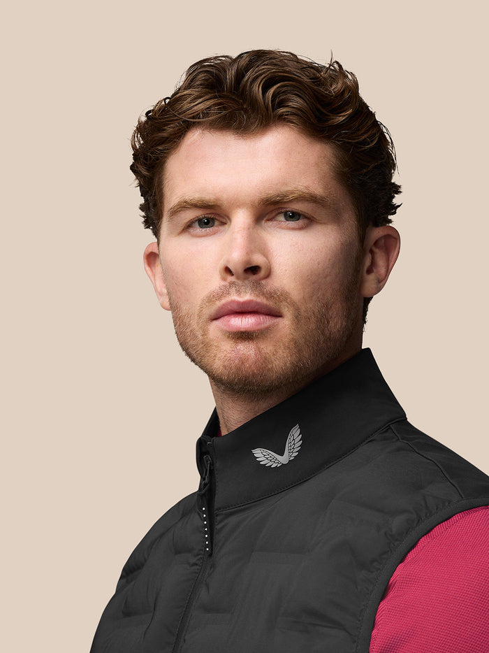 Side and back view of a man wearing a black Pinnacle Gilet over a pink short sleeve; Castore branding runs down the back.