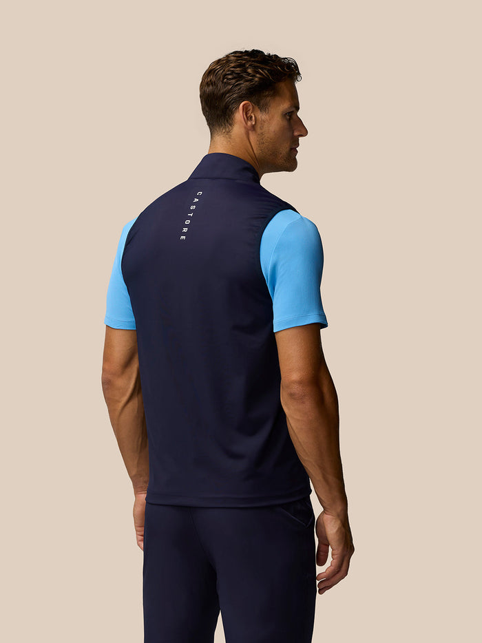 Back view of a man wearing a navy Pinnacle gilet with light blue sleeves and Castore branding along the upper back.