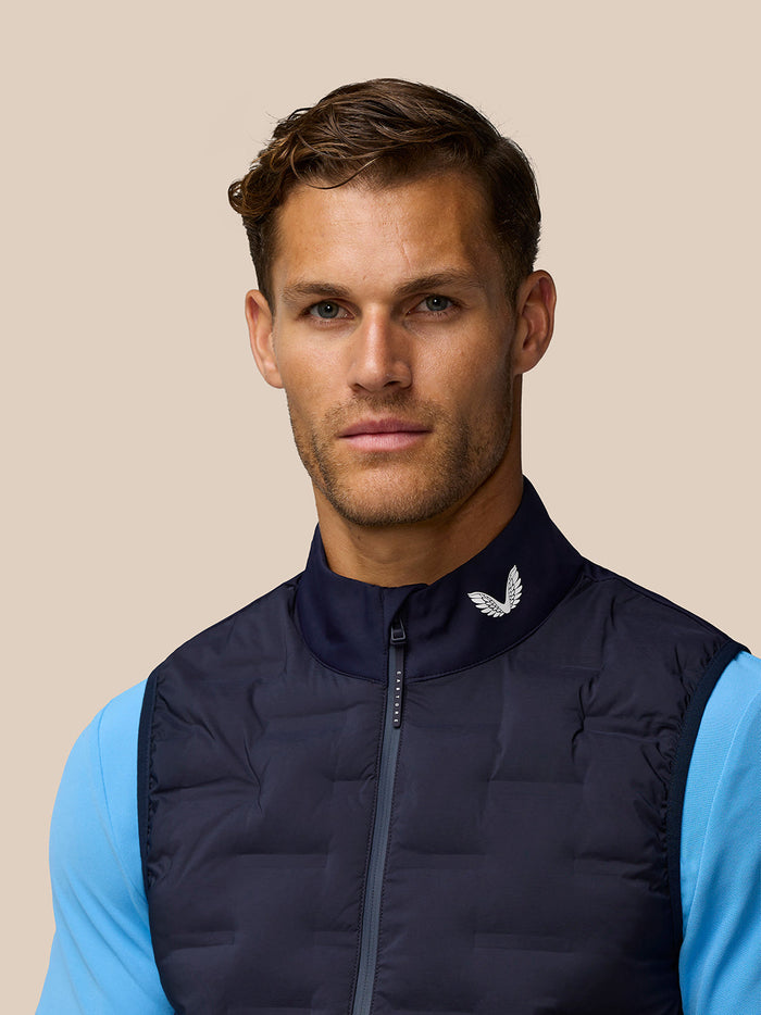 Close-up of a man in a navy quilted Pinnacle gilet with light blue sleeves and a white Castore logo on the collar.