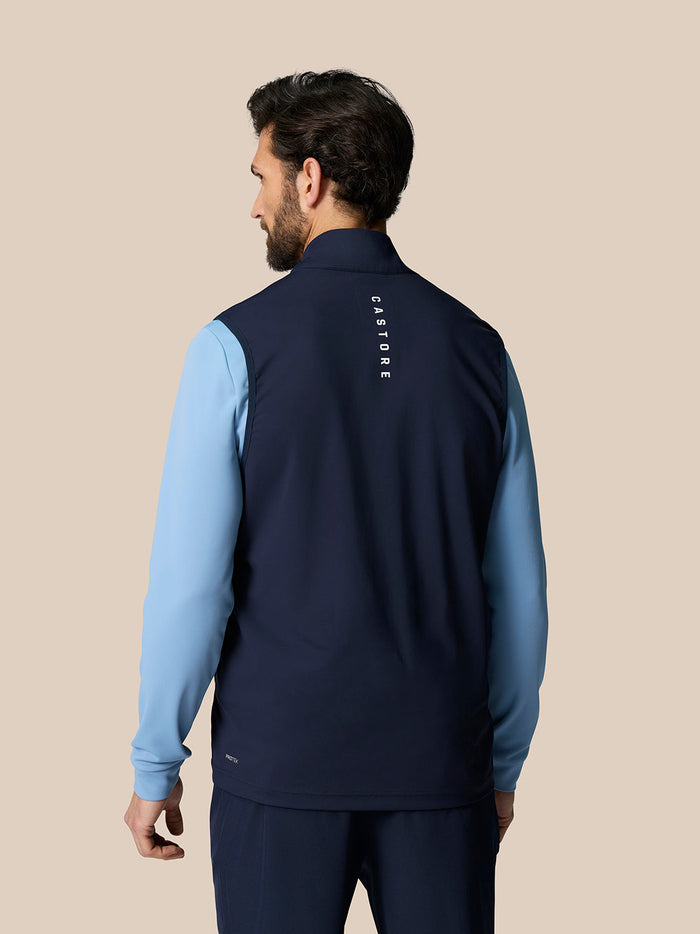 Back view of a man wearing a navy Pinnacle Gilet over a light blue long sleeve shirt, with Castore branding along the upper back