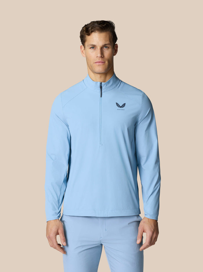 Front view of a man wearing a sky blue golf jacket with half zip and high funnel neckline, Castore logo on the chest.