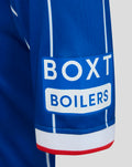 Sleeve detail of blue Rangers junior home shirt with BOX BOILERS sponsor patch and white cuff with red trim