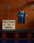 Blue Rangers FC home jersey hangs on a wooden locker wall beside a Welcome to Ibrox sign.