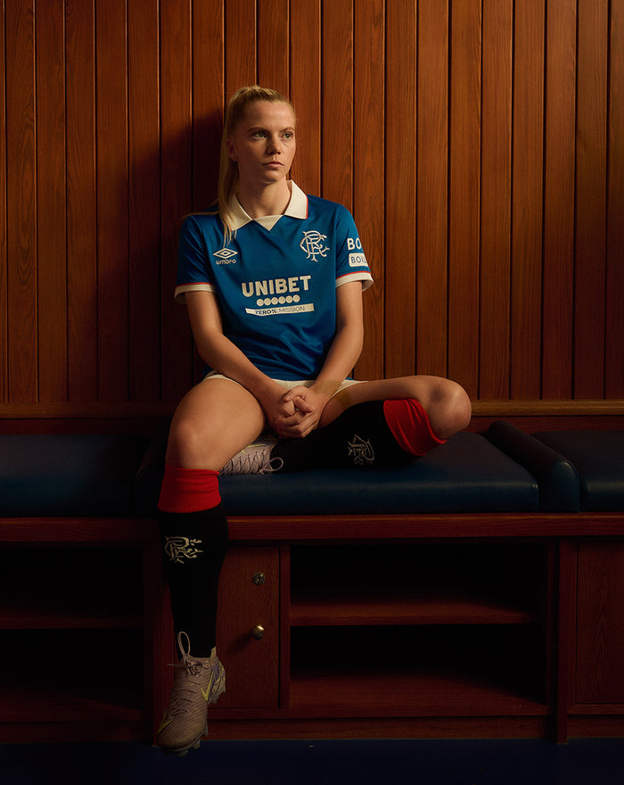 Woman seated on a wooden bench wearing a blue Rangers 25/26 Women’s Home Shirt with white collar Umbro logo club crest and UNIBET sponsor