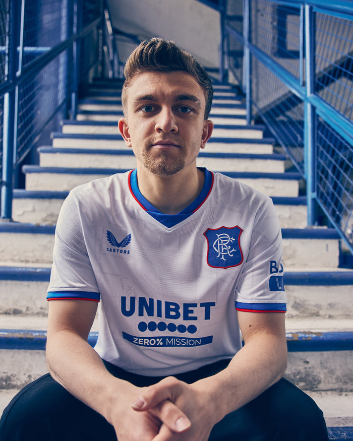 Portrait of a man wearing a white Rangers away shirt with crest and UNIBET sponsor, seated on blue stadium steps.