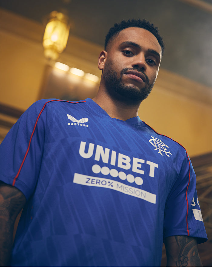 Man wearing blue Rangers Home Short Sleeve shirt with UNIBET sponsor and Rangers crest indoors.