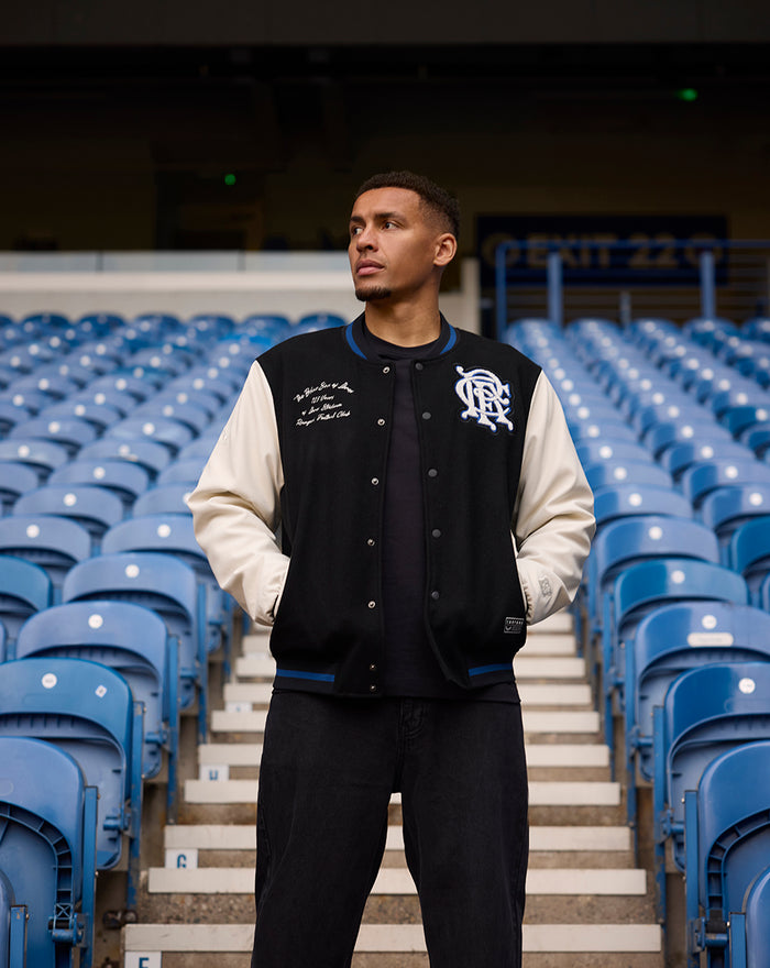 Model wearing black and cream Ibrox 125 Heritage Varsity Jacket with striped collar in stadium stands.