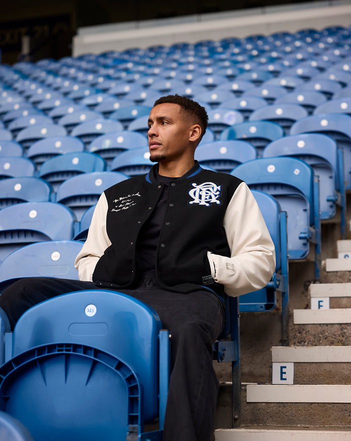 Man seated in stadium wearing black and cream Ibrox 125 Heritage Varsity Jacket with patches on chest.