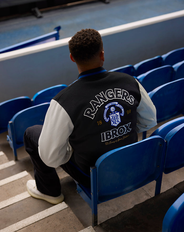 Back view of a man wearing a black and cream varsity jacket with a large Rangers Ibrox back graphic