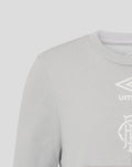 Close up of chest area on junior light gray T shirt showing Umbro logo and club crest.