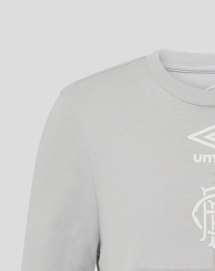 Close up of chest area on junior light gray T shirt showing Umbro logo and club crest.