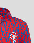 Close-up of a junior red and blue geometric print quarter zip jacket with a white club crest on the chest and a collar.