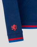 Close up of sleeve on blue hoodie showing a red crest logo near the cuff and a red blue striped ribbed cuff at the wrist