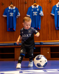 Toddler wearing Rangers FC Away 2025–26 infant third kit kicking a ball indoors with blue jerseys hanging in the background.