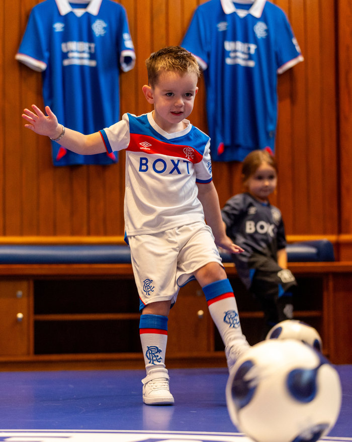 Young boy in Rangers infant away kit indoors kicks a soccer ball with another child in a dark kit in the background.