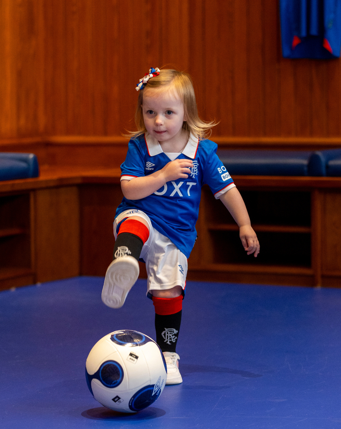 Toddler in Rangers FC infant 25/26 home kit with blue jersey white shorts red socks kicking a ball indoors