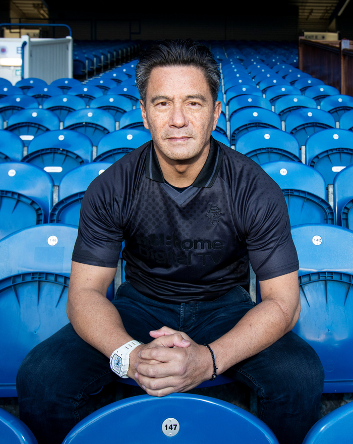 Man wearing a black UNISEX RETRO 2002-03 HOME BLACKOUT SHIRT seated among blue stadium seats