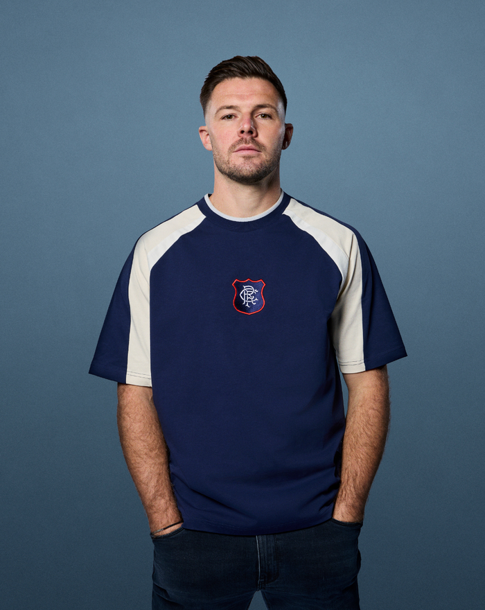 Male model wearing navy Rangers panelled 90s tee with cream shoulder panels against a blue backdrop.