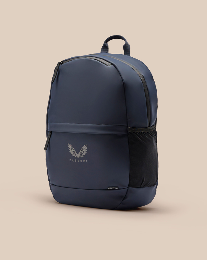 Blue CORE BACKPACK with top handle and side mesh pocket against a beige background featuring a front Castore logo