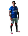 Model wearing blue Rangers junior home shirt with crest and sponsor, paired with white shorts and striped socks